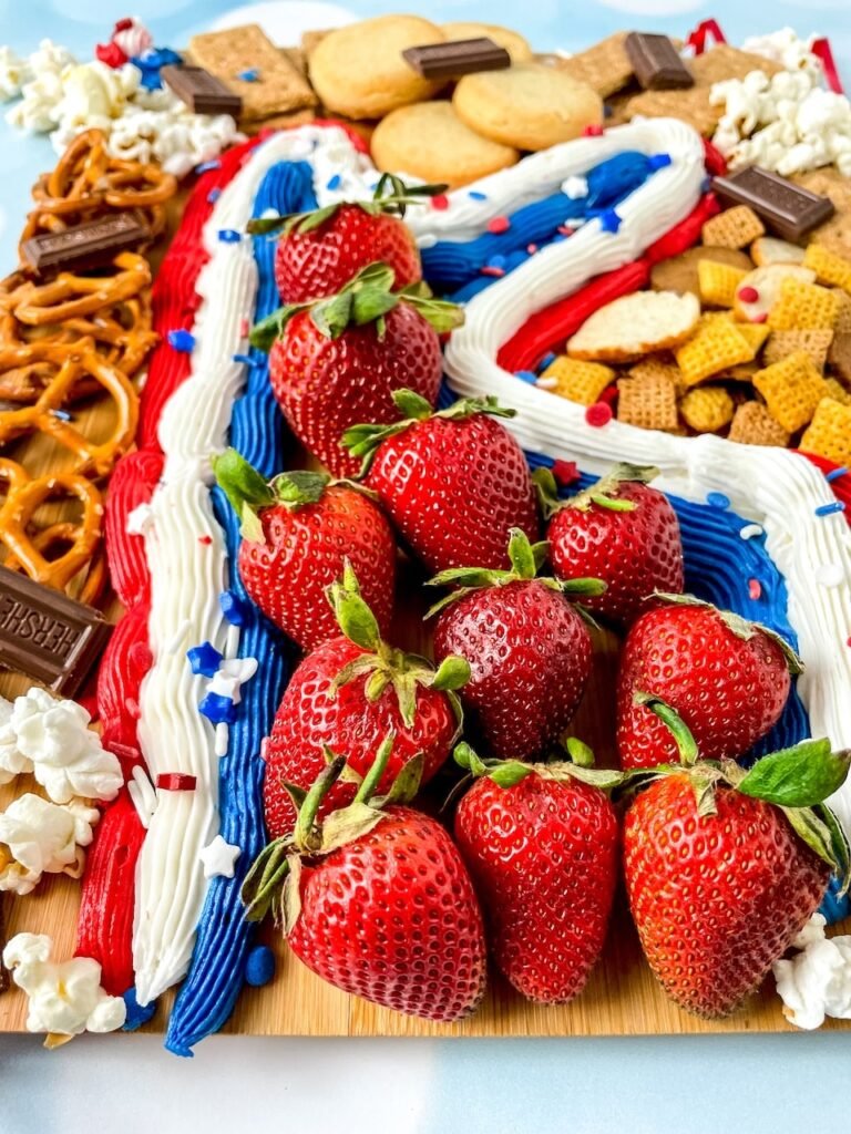 Close-up of a patriotic dessert board with fresh strawberries arranged over red, white, and blue buttercream frosting, surrounded by pretzels, cookies, popcorn, and chocolate pieces.