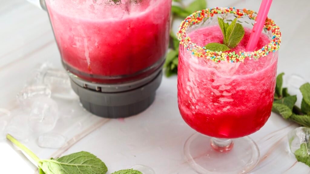 Glass of watermelon slushie with sprinkle rim and mint next to a blender filled with slushie, showing the finished drink and blending process