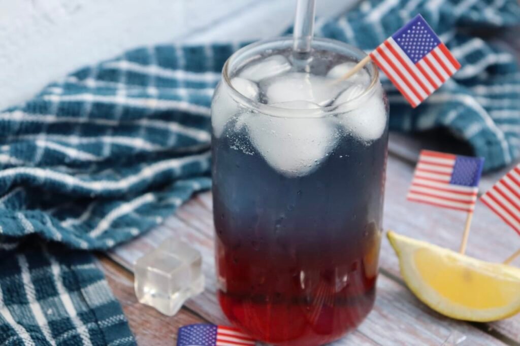 Close-up of a glass of patriotic punch with red, white, and blue layers, ice cubes, a straw, lemon wedge, and small American flags on a wooden table.