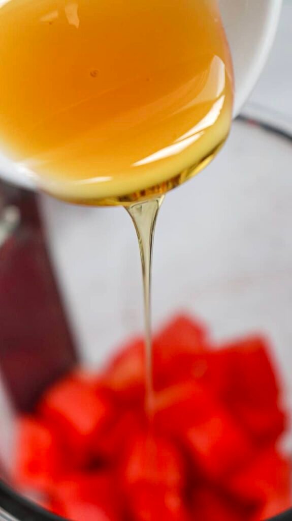 Close-up of honey being poured into a blender with cubed fruit, showing the step of adding sweetener to summer drink