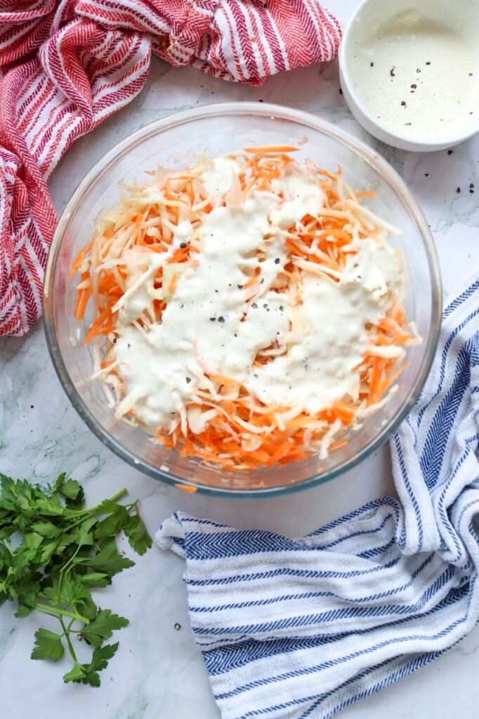 Overhead view of shredded cabbage and carrots in a bowl with creamy dressing poured on top, showing the step of combining ingredients.