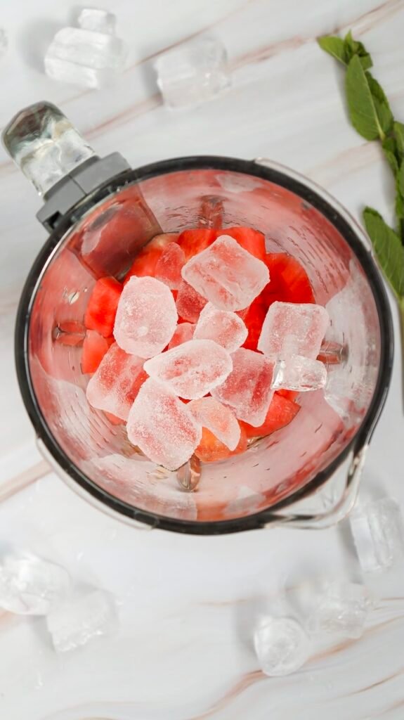 Top view of ice cubes added to cubed fruit in a blender, showing the step of creating a thick summer drink.