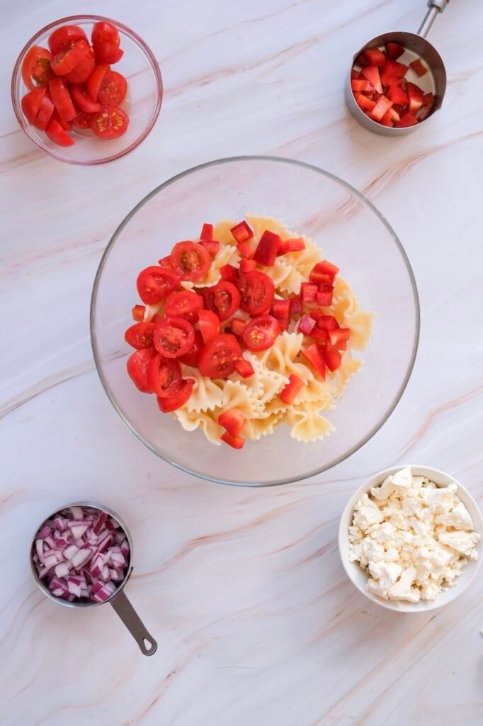 Overhead view of bowtie noodles in a glass bowl being topped with sliced cherry tomatoes and diced red bell pepper, with chopped red onion and feta cheese nearby during  preparation.