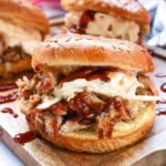Close-up of a BBQ pulled pork sandwich with shredded pork, barbecue sauce, and creamy coleslaw on a sesame seed bun, served on a wooden board.