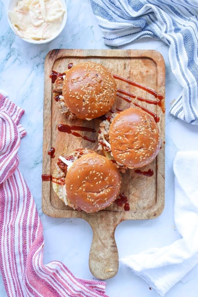 Overhead view of three BBQ pulled pork sandwiches on sesame seed buns with barbecue sauce on a wooden board, with coleslaw and kitchen towels nearby.