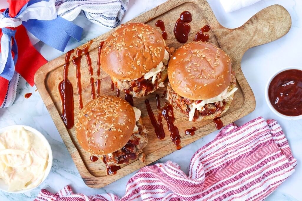 Overhead view of three BBQ pulled pork sandwiches with coleslaw on sesame seed buns, drizzled with barbecue sauce on a wooden board with side dishes, styled for a 4th of July cookout.