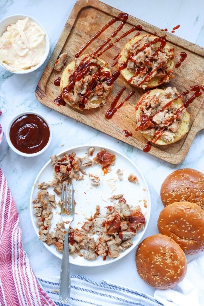 Open buns topped with shredded pig meat and sauce on a wooden board, with extra pork, buns, and coleslaw nearby, showing how to assemble meal.