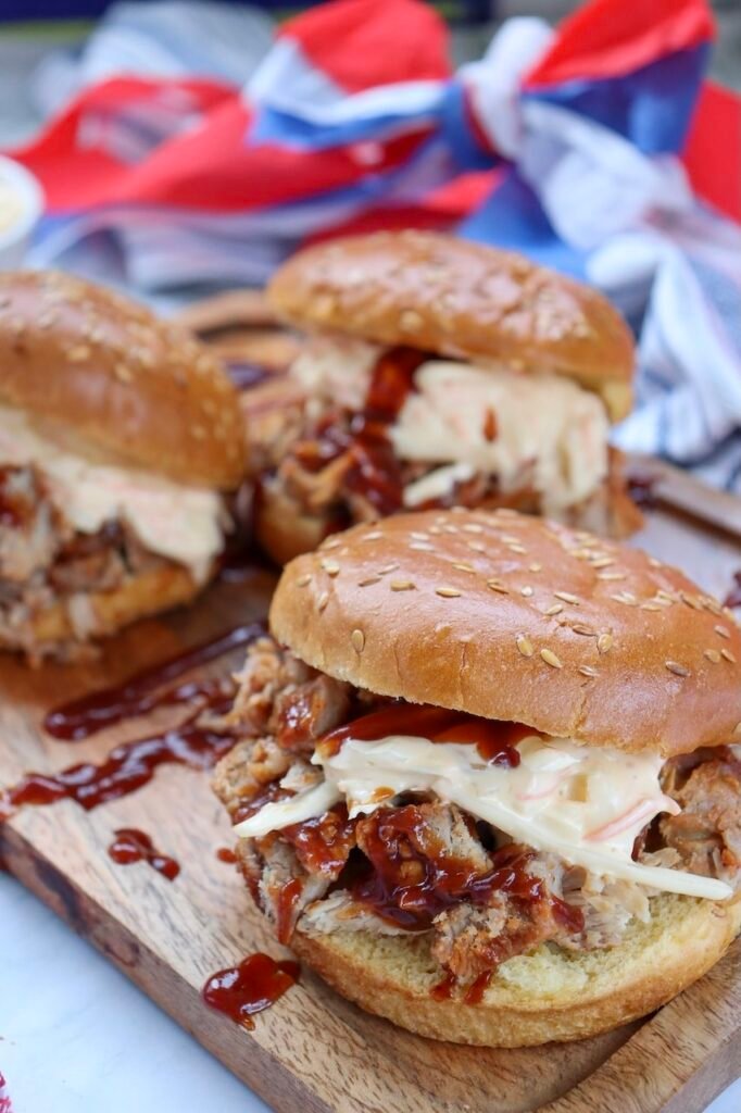 Close-up of BBQ pulled pork sandwiches with coleslaw on sesame seed buns, served on a wooden board with barbecue sauce and red, white, and blue cloth in the background.