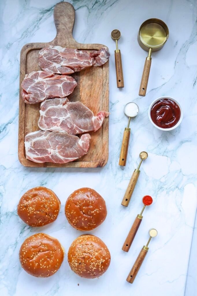 Raw pork shoulder pieces on a wooden cutting board with BBQ sauce, spices, oil, and sandwich buns arranged on a marble surface, showing the ingredients needed to make BBQ pulled pork sandwiches.