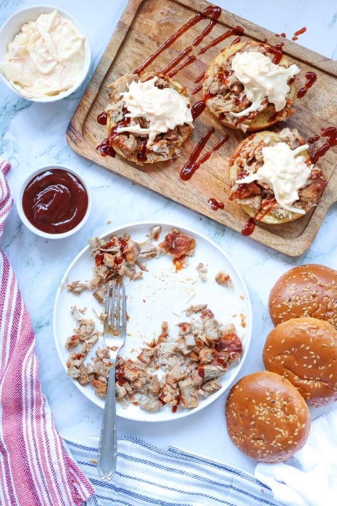 Open buns topped with shredded pig meat, sauce, and creamy coleslaw on a wooden board, with extra pork, buns, and sauce nearby, showing the final assembled food.
