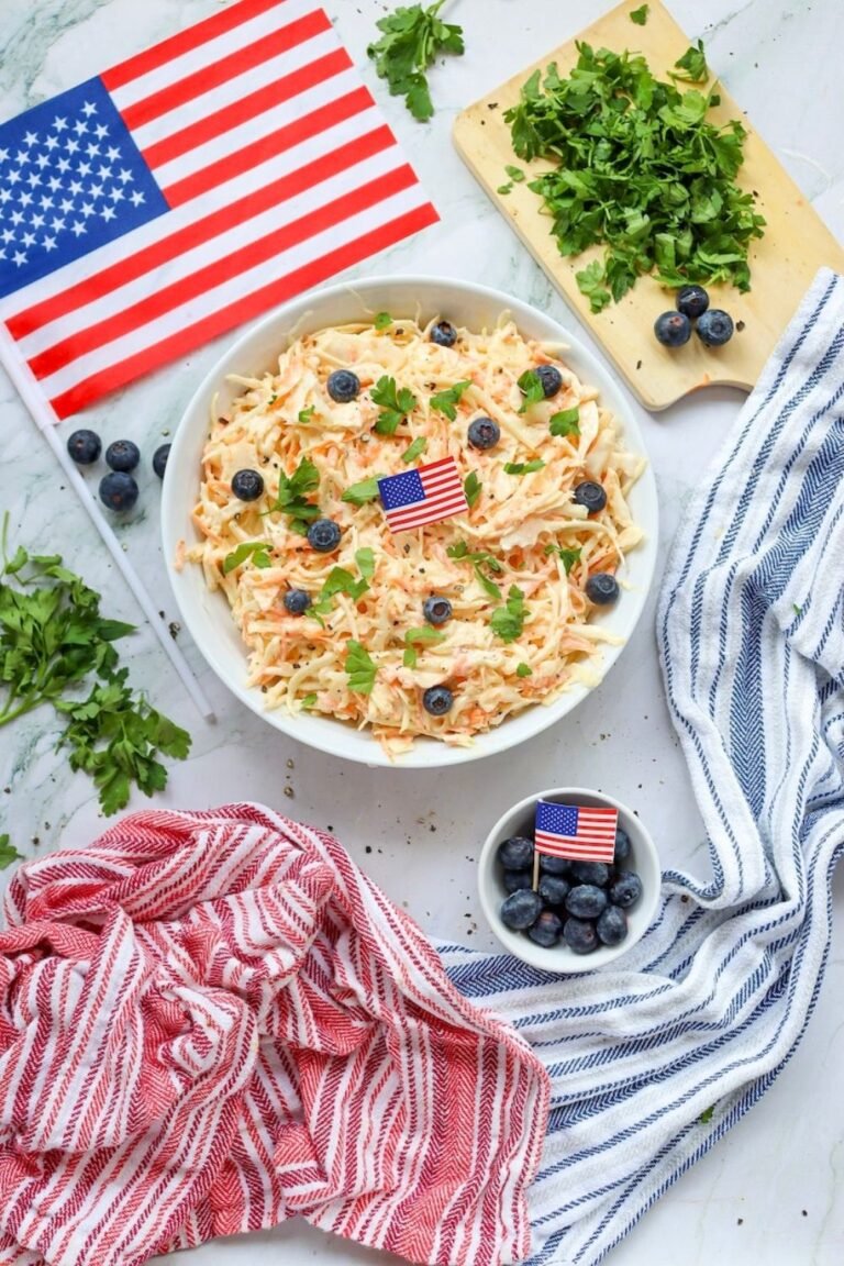 Overhead view of a bowl of creamy coleslaw with shredded cabbage and carrots, topped with parsley and blueberries, styled with an American flag for a 4th of July presentation.