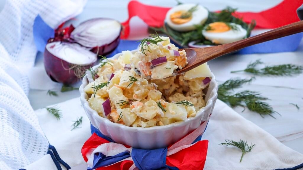 Spoon lifting a serving of creamy potato salad with potatoes, eggs, red onion, and dill from a bowl decorated with red, white, and blue for a Fourth of July meal.