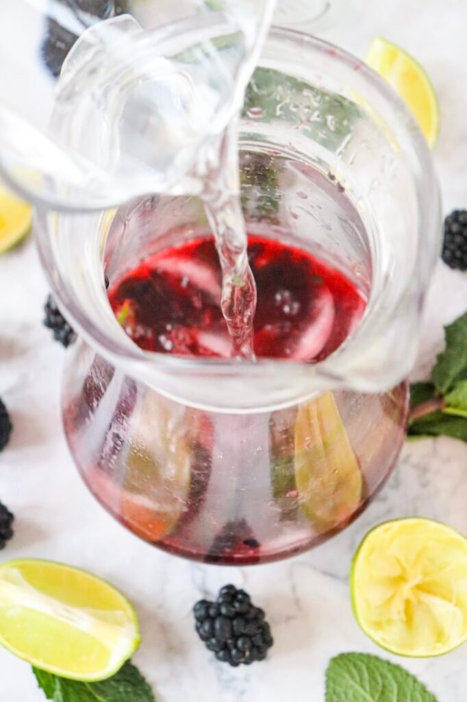 Sparkling water being poured into a blackberry mojito mocktail mixture with ice, lime, and mint in a glass pitcher
