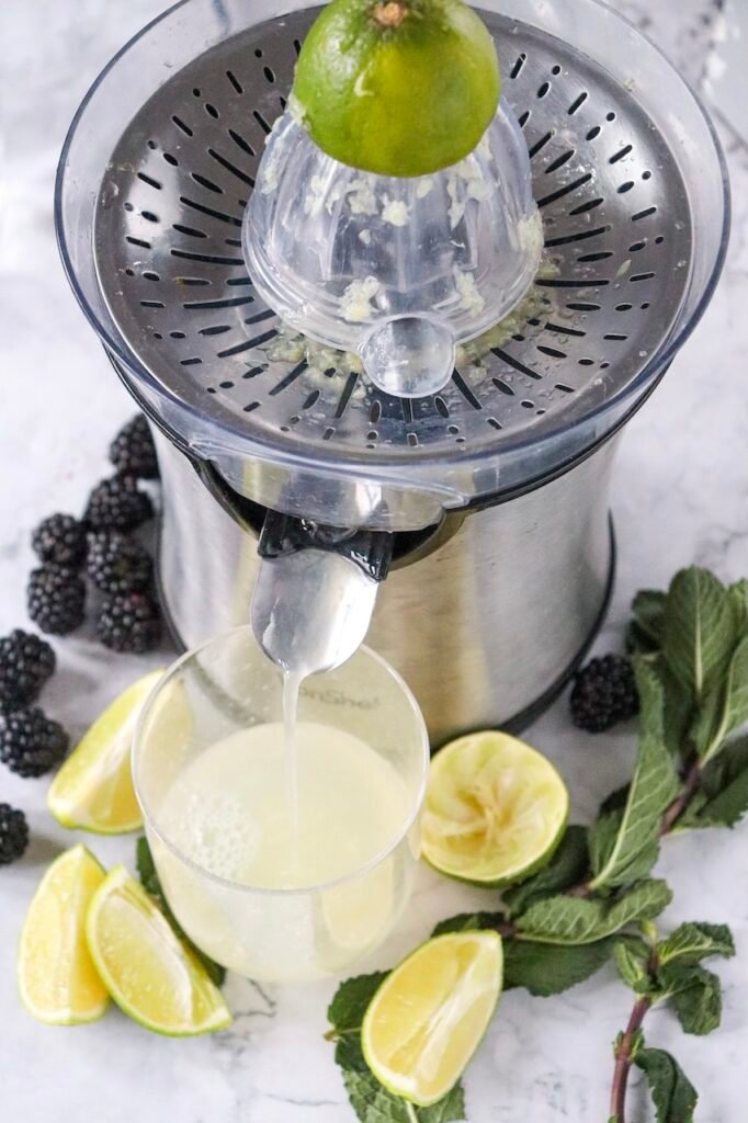 Fresh lime being squeezed with a juicer into a glass while preparing a blackberry mojito mocktail with blackberries and mint nearby