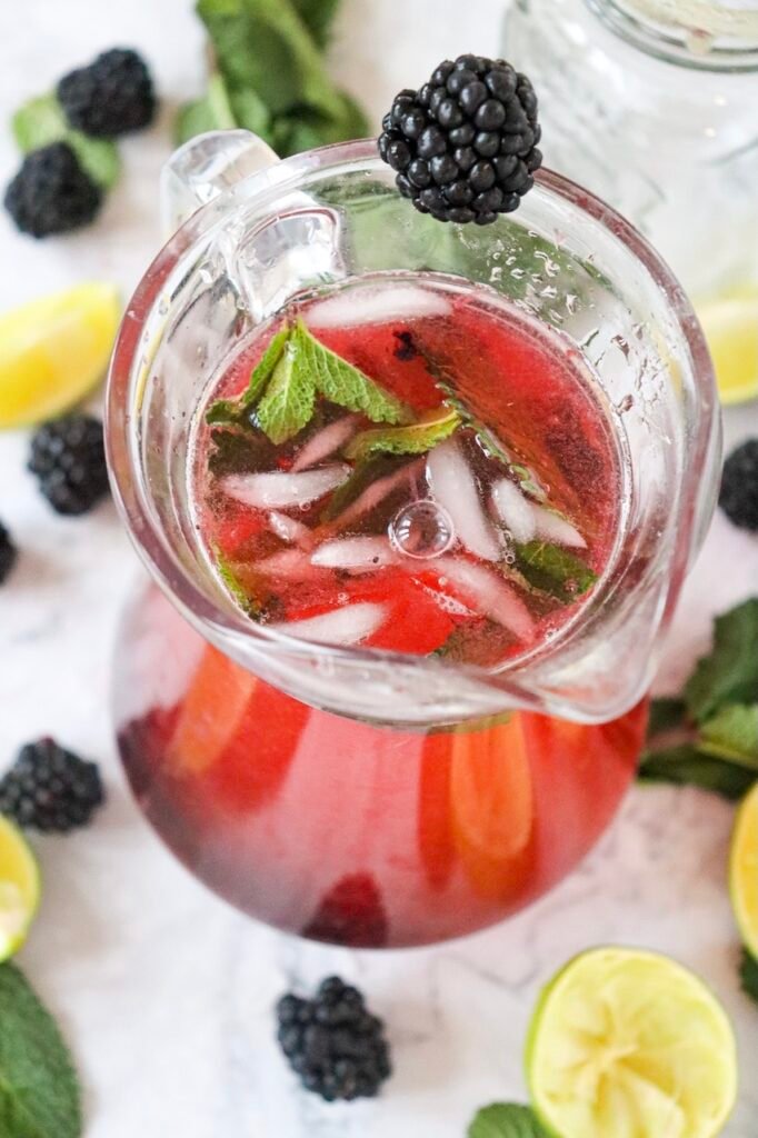 Glass pitcher filled with a blackberry mojito mocktail with ice, mint leaves, and a blackberry garnish, surrounded by lime wedges and fresh mint