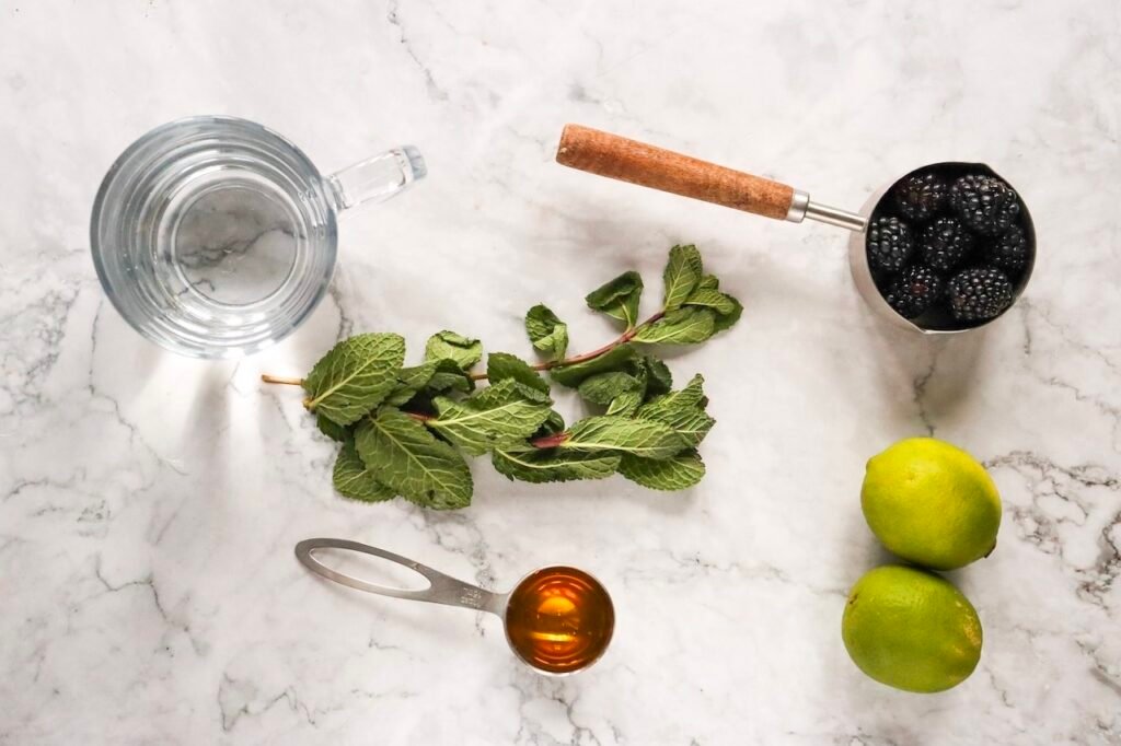 Fresh ingredients for a blackberry mojito mocktail including blackberries, mint leaves, limes, agave syrup, and sparkling water arranged on a marble surface