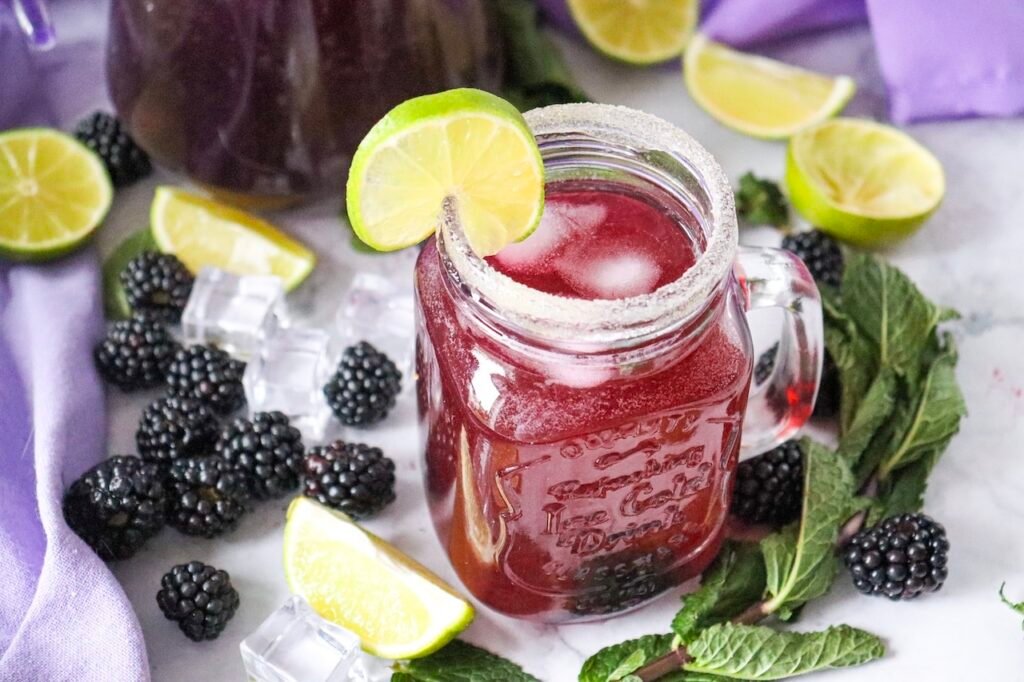 Mason jar filled with a blackberry mojito mocktail with ice, mint, lime slice garnish, and sugared rim surrounded by blackberries and mint