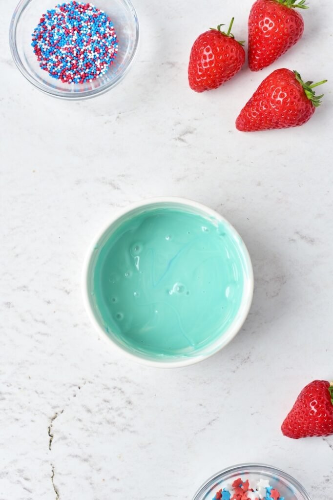 Light blue melted chocolate in a small bowl with fresh strawberries and colorful sprinkles nearby for decorating a patriotic dessert.