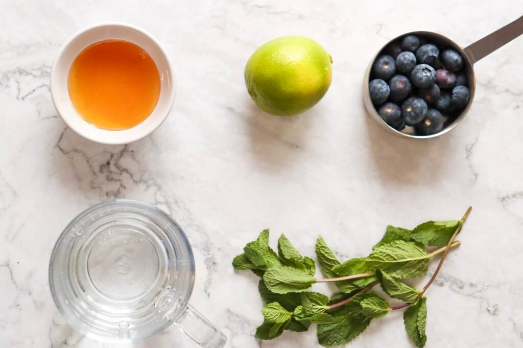 Fresh ingredients for a blueberry mojito mocktail including blueberries, mint leaves, lime, honey, and sparkling water arranged on a marble surface.