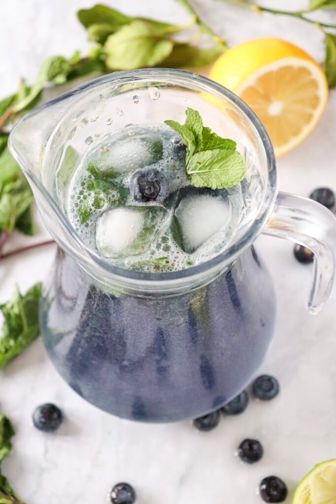 Glass pitcher filled with blueberry mojito mocktail, ice, mint leaves, and fresh blueberries, with lime and mint in the background.