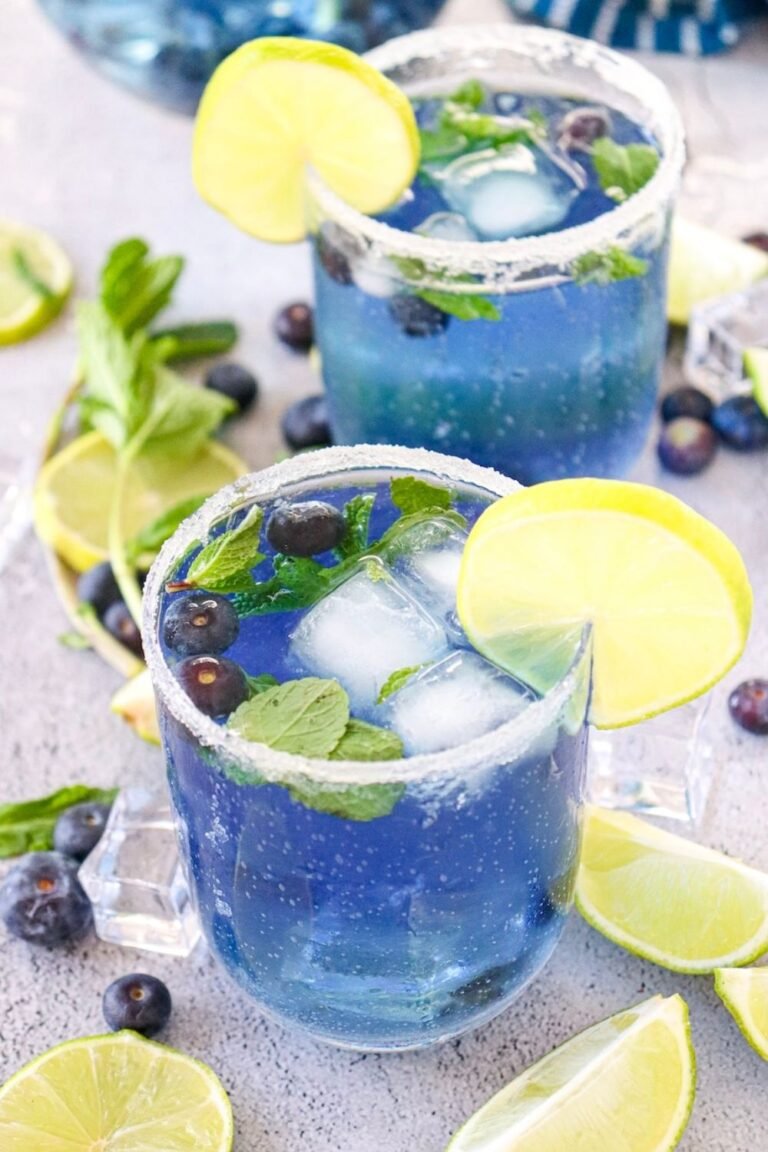 Two glasses of blueberry mojito mocktail with ice, mint leaves, blueberries, and lime slices, served with a sugared rim on a light surface.