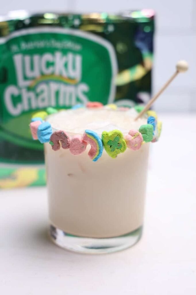 Creamy boozy Lucky Charms drink in a glass with ice and a marshmallow-covered rim, with cereal in the background.