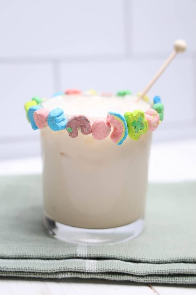 Creamy boozy Lucky Charms drink in a glass with a colorful marshmallow rim and stir stick on a cloth napkin.
