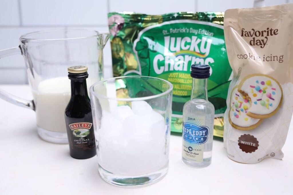 Glass filled with ice surrounded by Baileys Irish Cream, vodka, milk, Lucky Charms cereal, and cookie icing, showing ingredients for a boozy Lucky Charms drink.