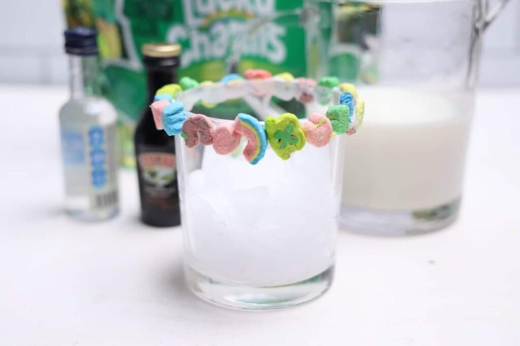 Glass filled with ice and rimmed with white icing and cereal marshmallows, with drink ingredients blurred in the background.