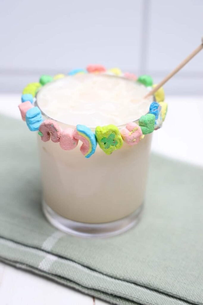 Creamy boozy Lucky Charms drink in a glass with a colorful marshmallow rim and stir stick, set on a cloth napkin.