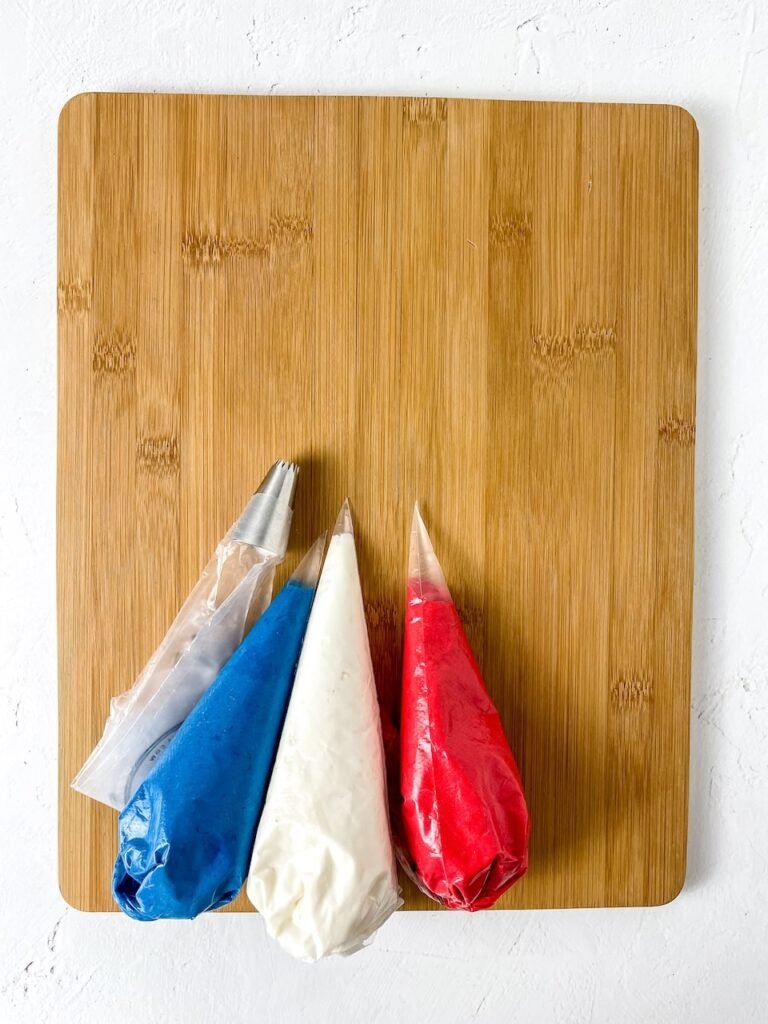 Four piping bags filled with red, white, and blue buttercream frosting resting on a wooden board, showing the setup step before decorating.