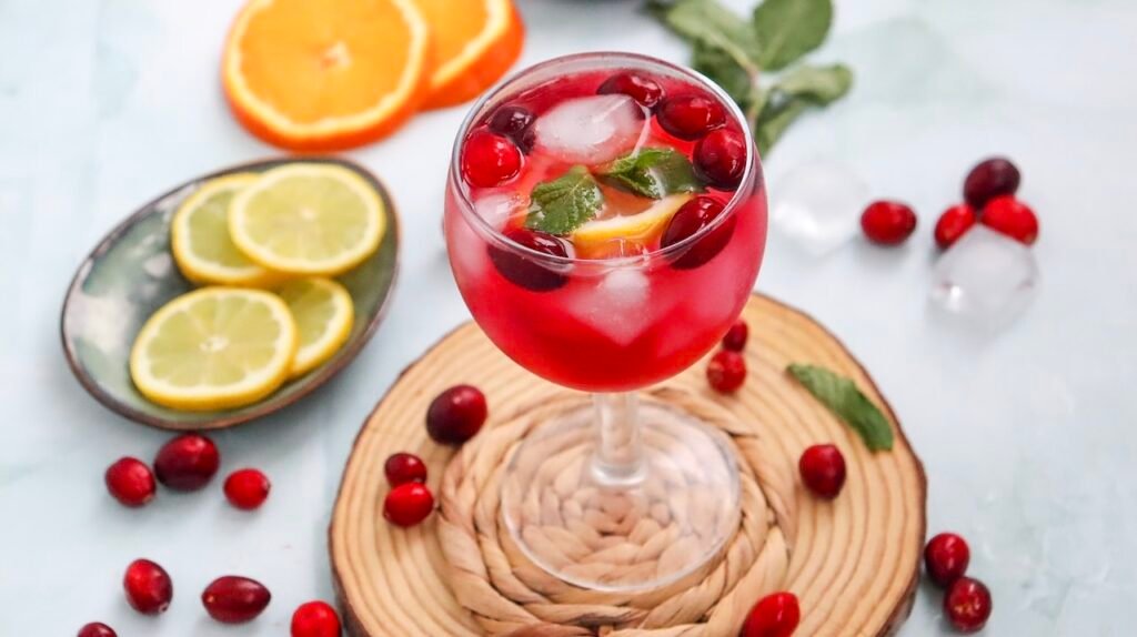 Wide shot of a cranberry spritzer mocktail in a glass with ice, garnished with cranberries, citrus slices, and mint, surrounded by fresh ingredients on a light surface.