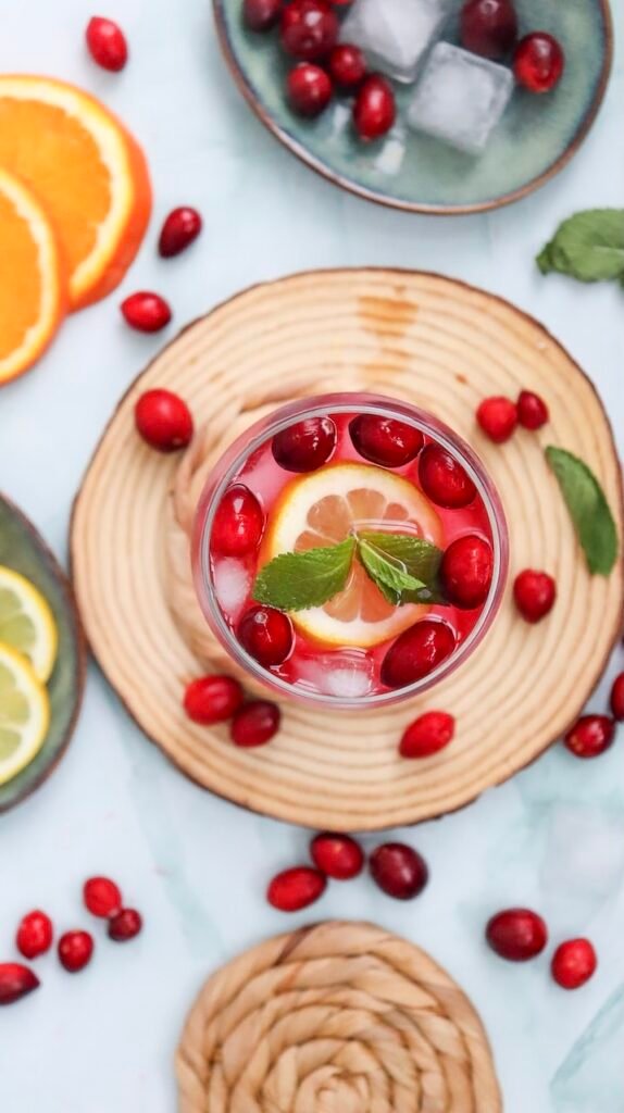 Top view of a cranberry spritzer mocktail in a glass with ice, garnished with cranberries, citrus slices, and mint, surrounded by fresh ingredients on a light surface.