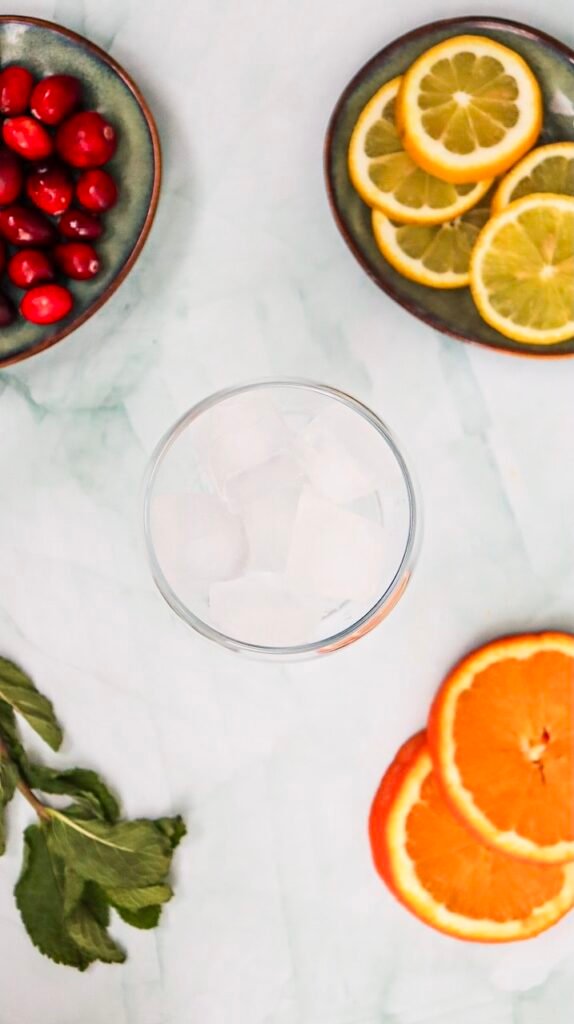 Overhead view of a glass filled with ice cubes surrounded by cranberries, citrus slices, and mint, showing the first step of preparing a drink.
