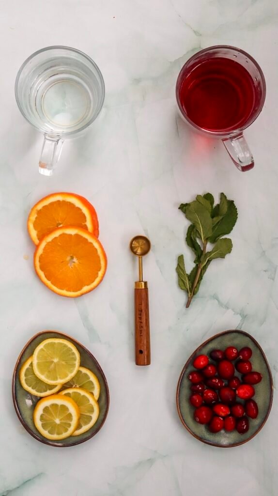 Flat lay of ingredients for a cranberry spritzer mocktail, including cranberry juice, sparkling water, sliced oranges and lemons, fresh cranberries, mint leaves, and a measuring spoon on a light marble surface.