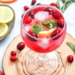 Glass of cranberry spritzer mocktail filled with ice and garnished with fresh cranberries, citrus slices, and mint, served on a wooden coaster.