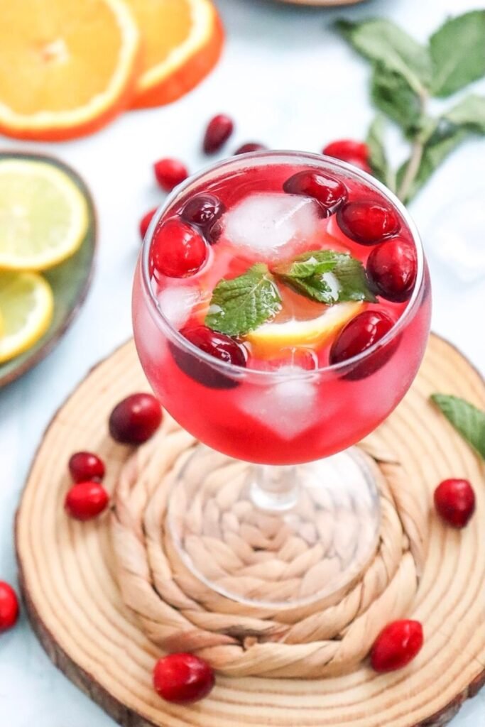 Glass of cranberry spritzer mocktail filled with ice and garnished with fresh cranberries, citrus slices, and mint, served on a wooden coaster.