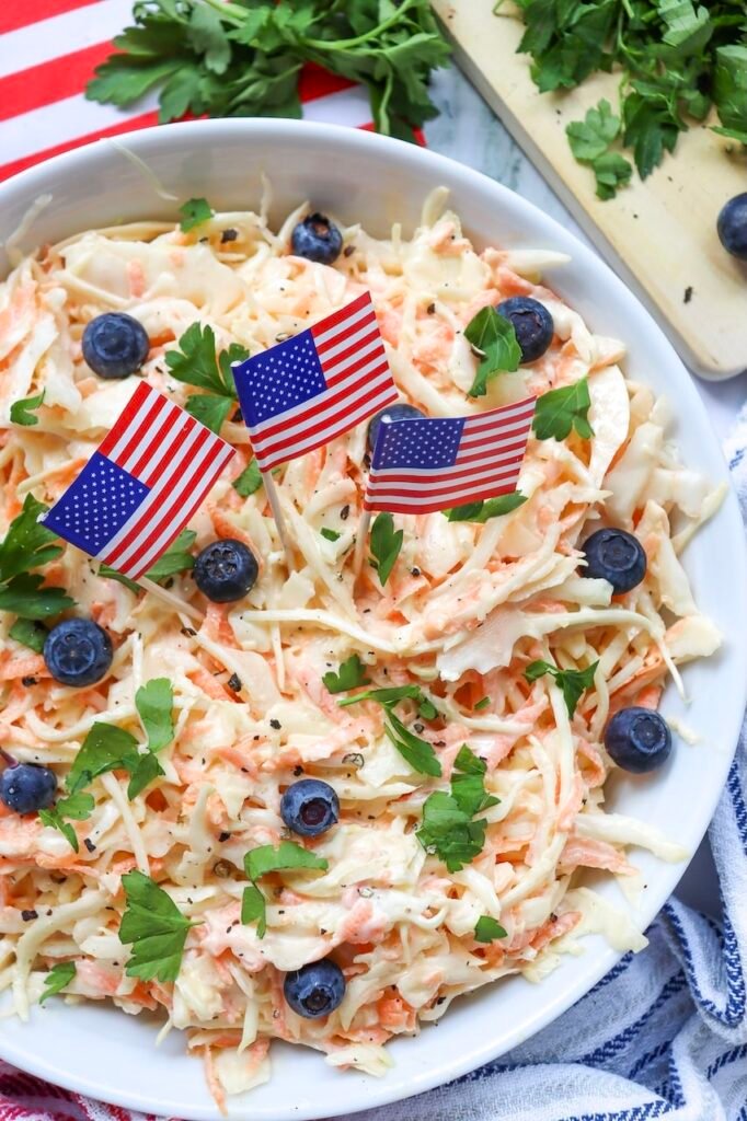 Close-up view of creamy coleslaw with shredded cabbage and carrots, topped with blueberries, parsley, and small American flags for a festive 4th of July presentation.
