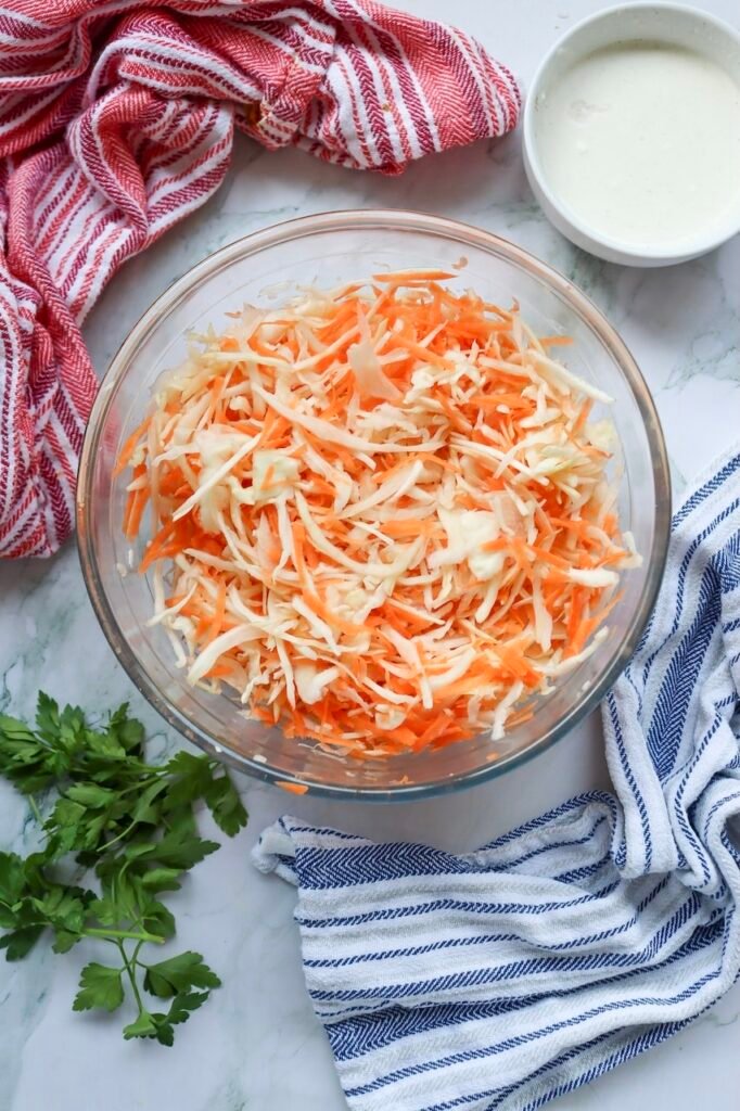 Overhead view of a bowl of freshly mixed cabbage with shredded cabbage and carrots, surrounded by kitchen towels and dressing on a marble surface.