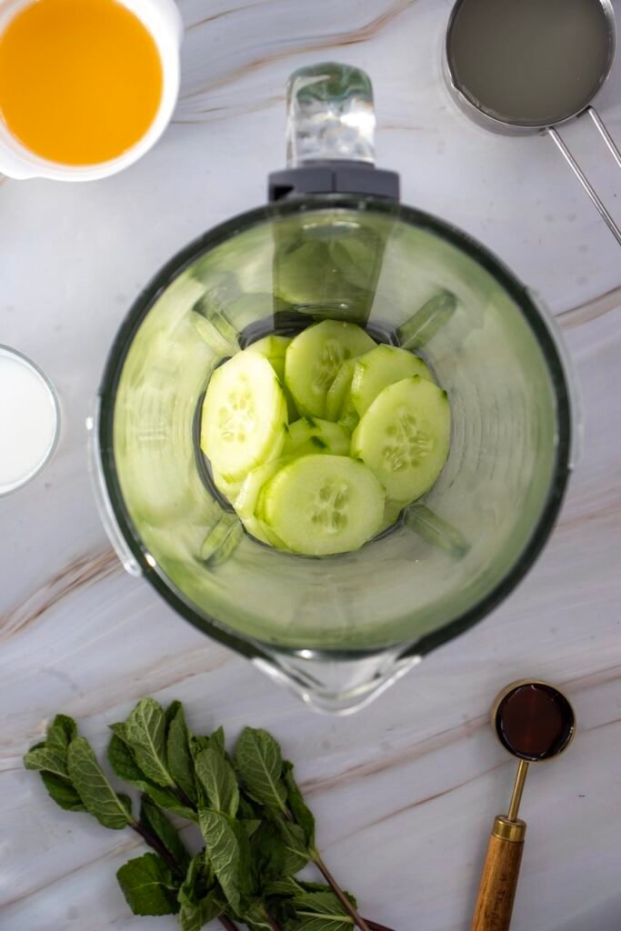 Top-down view of cucumber margarita mocktail ingredients including sliced cucumber in a bowl, lime juice, orange juice, agave, and fresh mint arranged on a light marble surface.