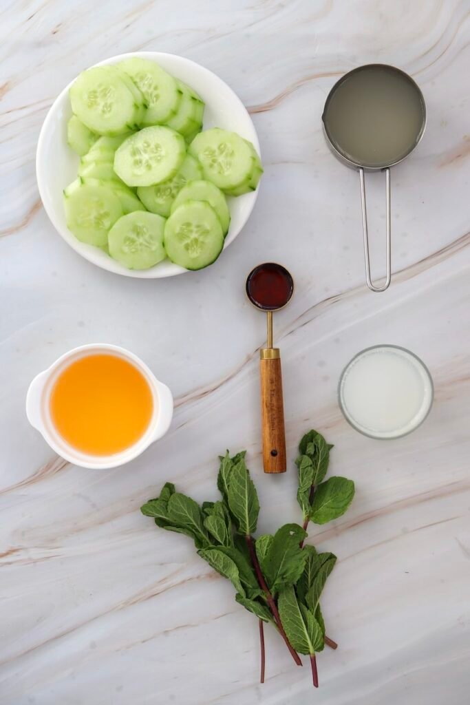 Top-down view of cucumber margarita mocktail ingredients including sliced cucumber in a bowl, lime juice, orange juice, agave, and fresh mint arranged on a light marble surface.