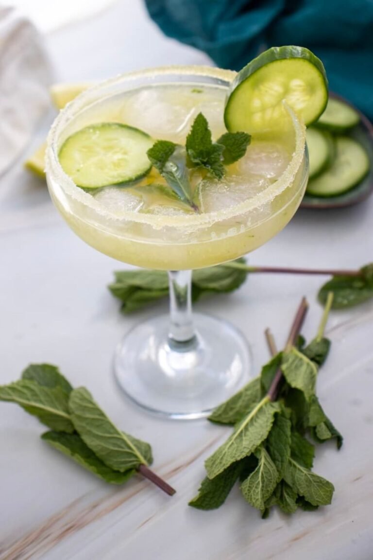 Cucumber margarita mocktail served in a glass with a salted rim, ice, cucumber slices, and fresh mint garnish on a light surface.