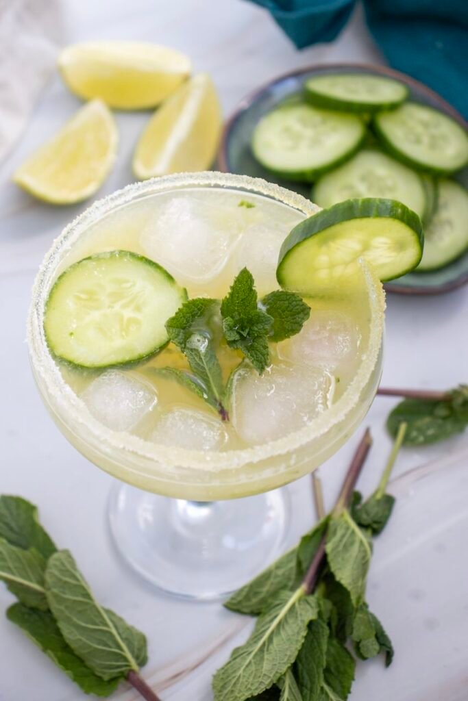 Top-down view of a cucumber margarita mocktail with a salted rim, ice, cucumber slices, and mint garnish, with lime wedges and sliced cucumber in the background.