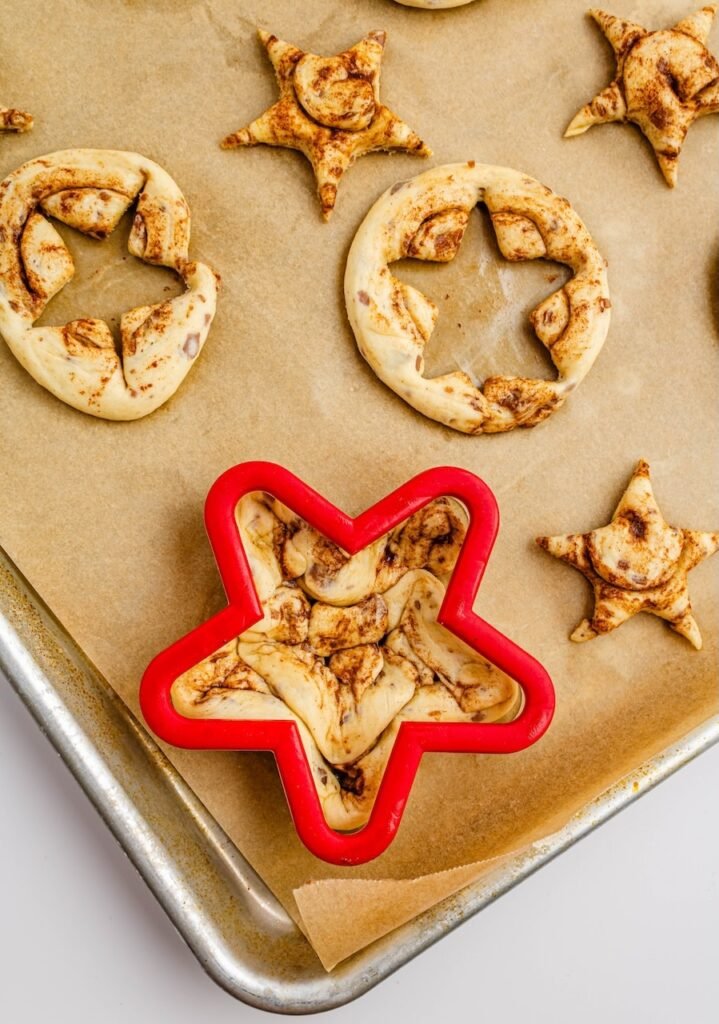 Overhead view of cinnamon roll dough being pressed into a red star-shaped cookie cutter on a parchment-lined baking sheet, with cut star shapes and dough scraps nearby.
