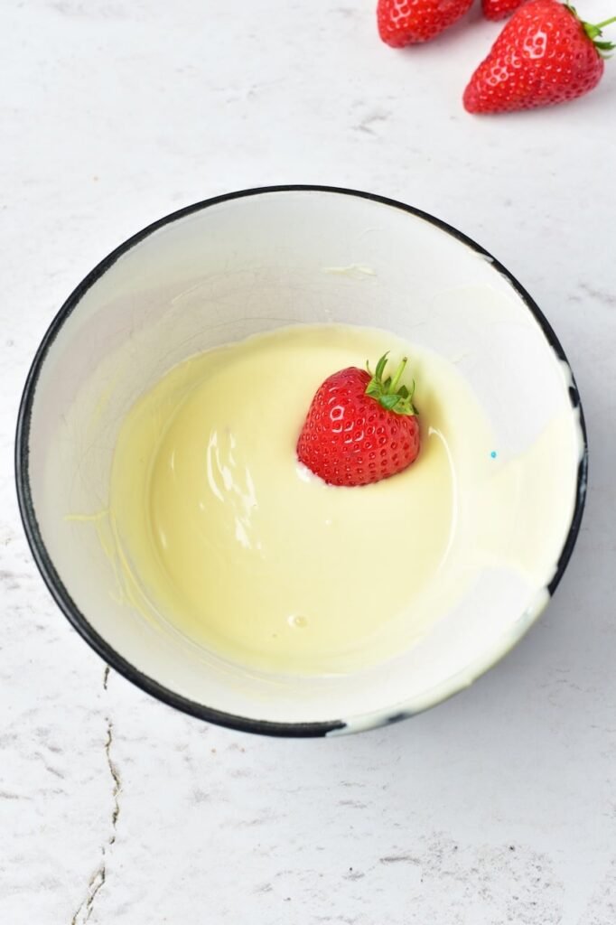 A fresh strawberry being dipped into melted white chocolate in a bowl as part of making a patriotic dessert.