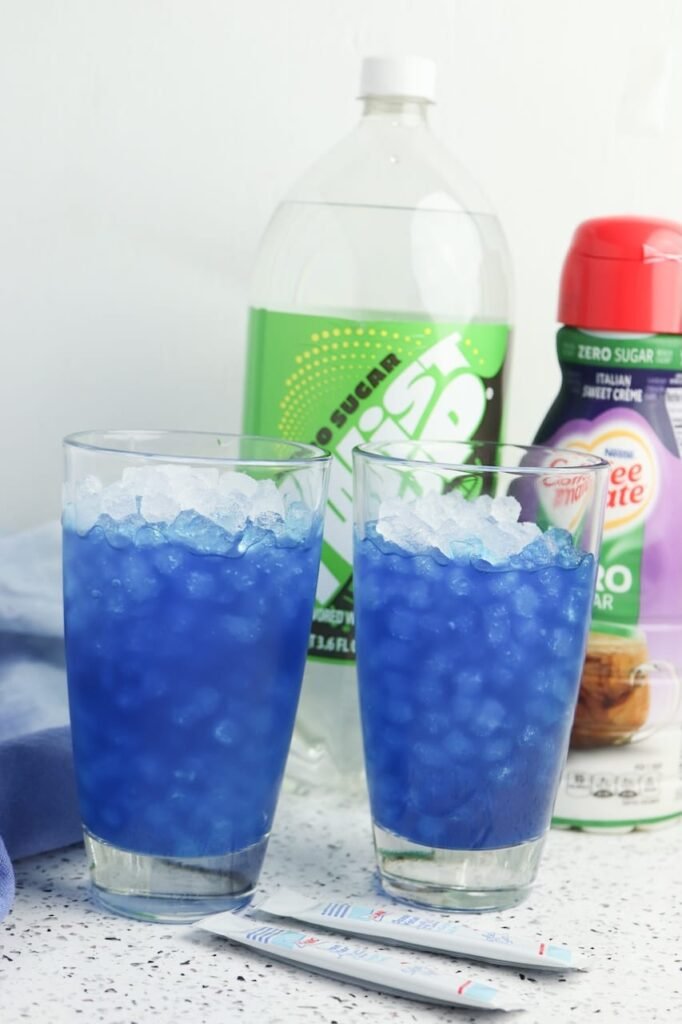 Two glasses filled with bright blue Dirty Ocean Water and crushed ice, with soda and coffee creamer in the background
