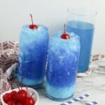 Two glasses of Dirty Ocean Water with creamy swirl and crushed ice, topped with cherries, with extra cherries and striped straws on the table