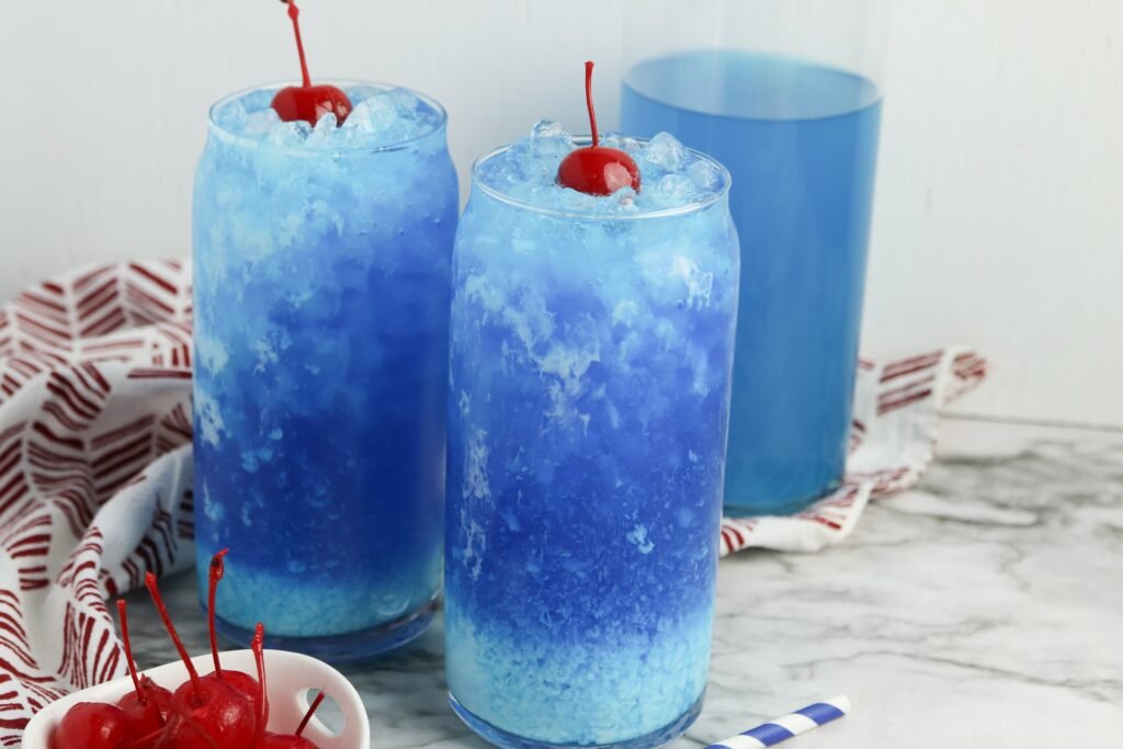 Two tall glasses of filled with crushed ice and a creamy blue swirl, each topped with a bright red cherry, with extra cherries in a bowl and a glass of blue drink in the background