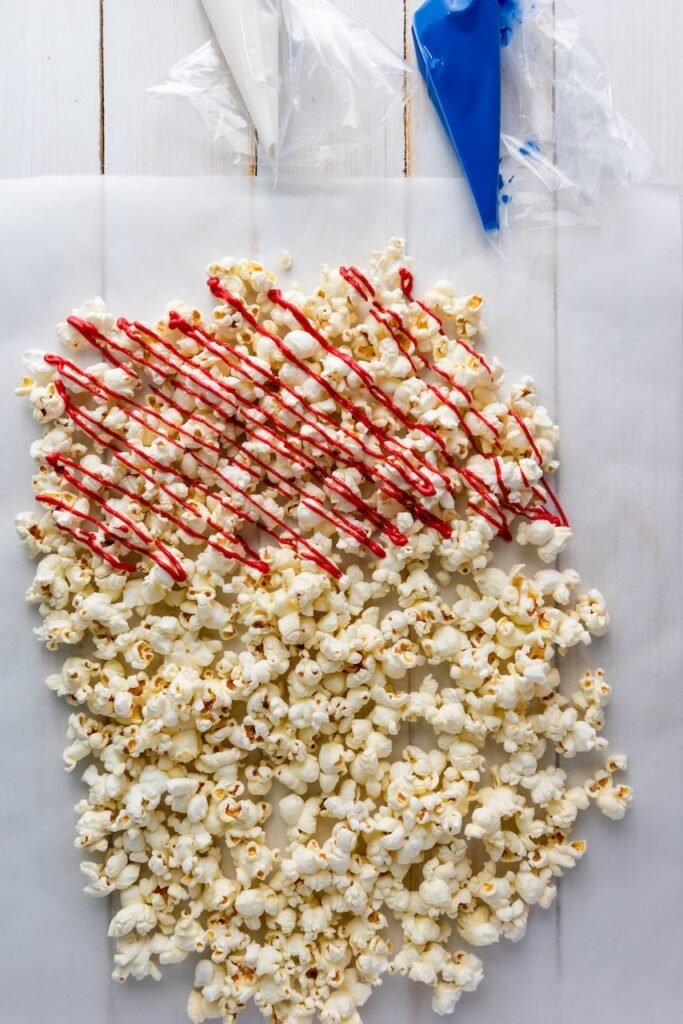 Freshly popped popcorn spread on parchment paper with red candy melts being drizzled over the top, showing a step in making 4th of July popcorn.