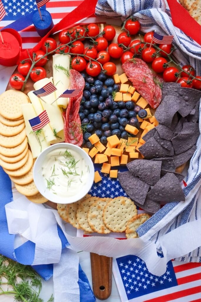 Vertical image of a 4th of July charcuterie board on a wooden platter with blueberries in the center, cherry tomatoes on the vine, cheddar cubes, Swiss cheese slices, crackers, blue tortilla chips, salami, and a bowl of dip, decorated with small American flags and patriotic cloth.